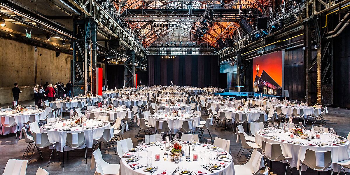 Große, für eine formelle Veranstaltung eingerichtete Industriehalle mit zahlreichen runden Tischen mit weißen Tischdecken, eleganten Gedecken und Tafelaufsätzen, umgeben von weißen Stühlen unter einer hohen, freiliegenden Stahldecke.